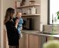 Preview: Eine Frau mit Baby steht in einer Küche. Im Hintergrund steht dezent ein weißer Luftentfeuchter.